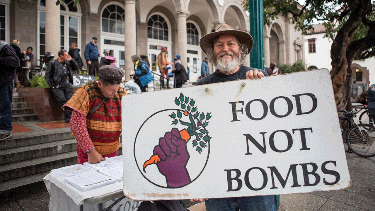 Keith McHenry holds the weathered Food Not Bombs sign in front of the the Santa Cruz Post Office and smiles.